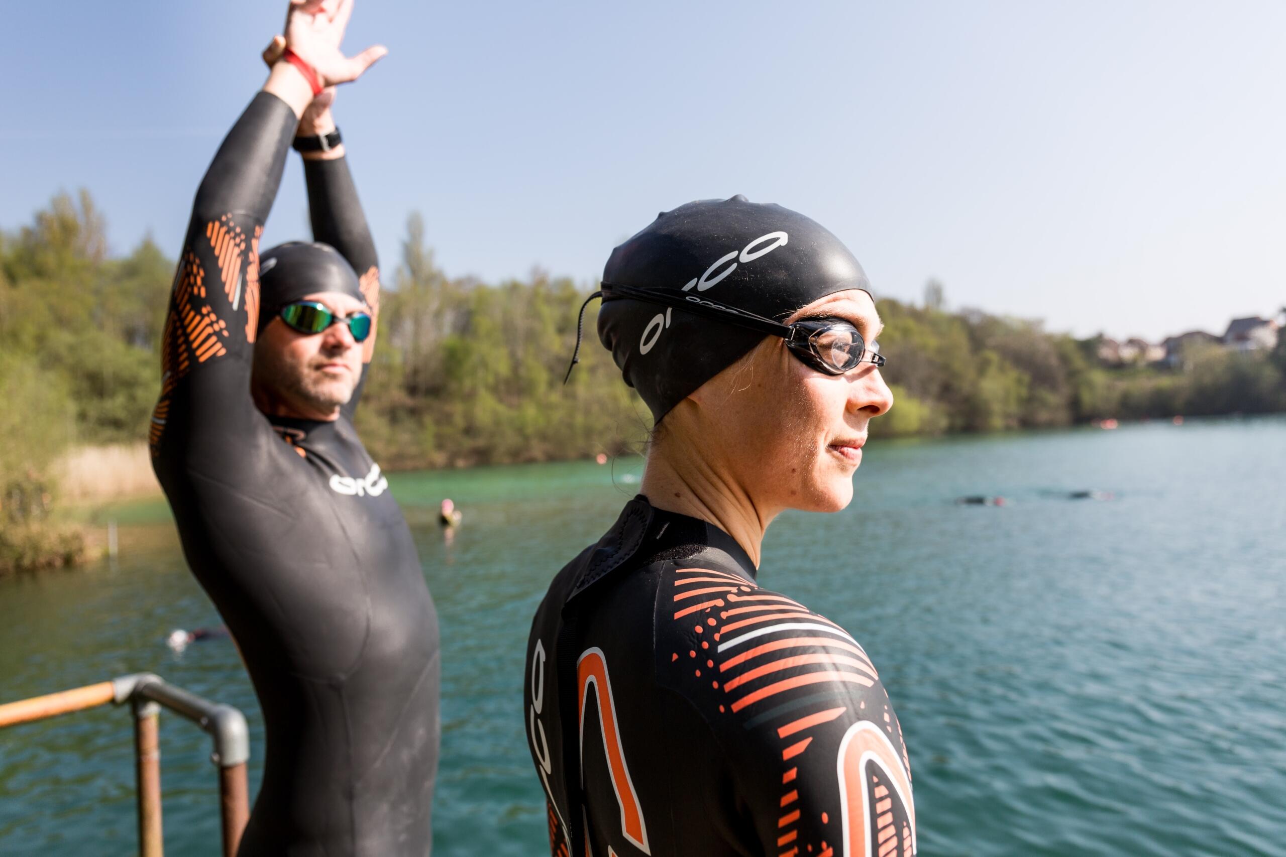 woman and man in wetsuits, goggles and swimming caps looking across a lake