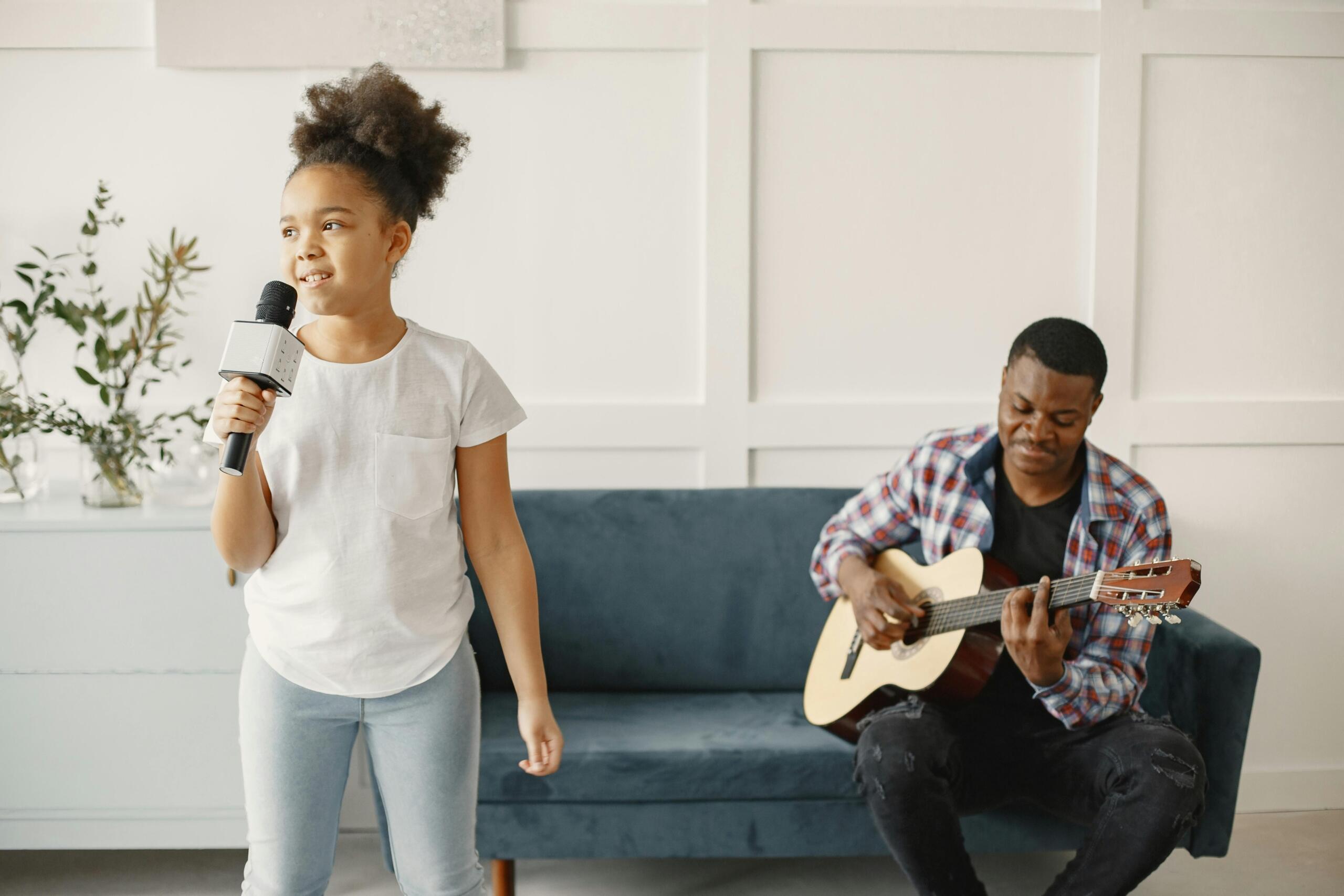 girls singing into wireless microphone while her teacher sits on sofa playing guitar