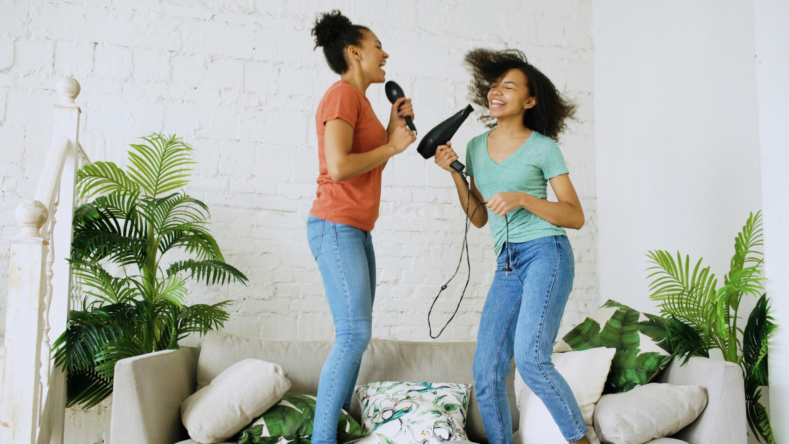 two girls dancing on a sofa singing into a hairbrush and hairdryer