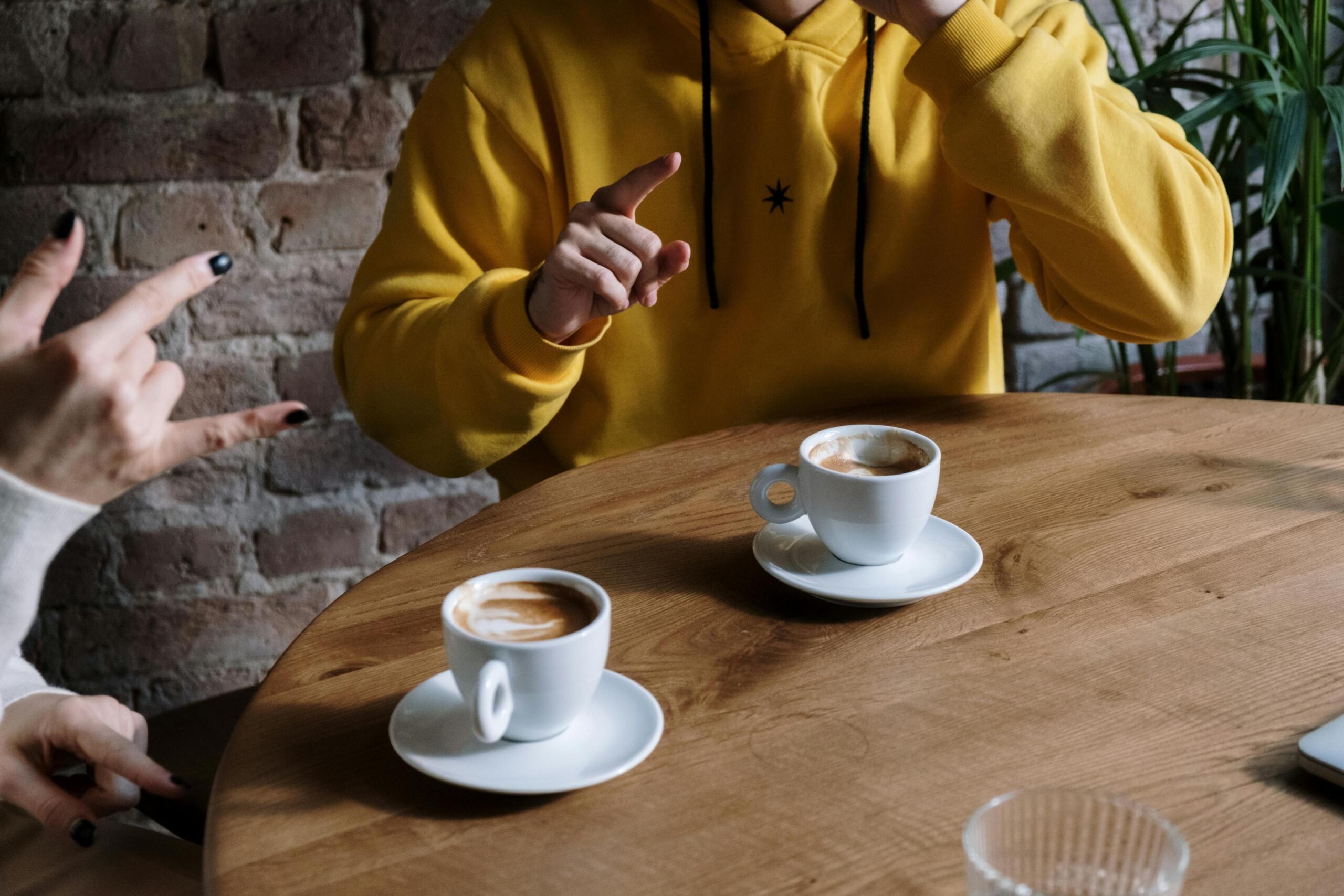 two people sitting across from each other at a table drinking coffee and using sign language gestures