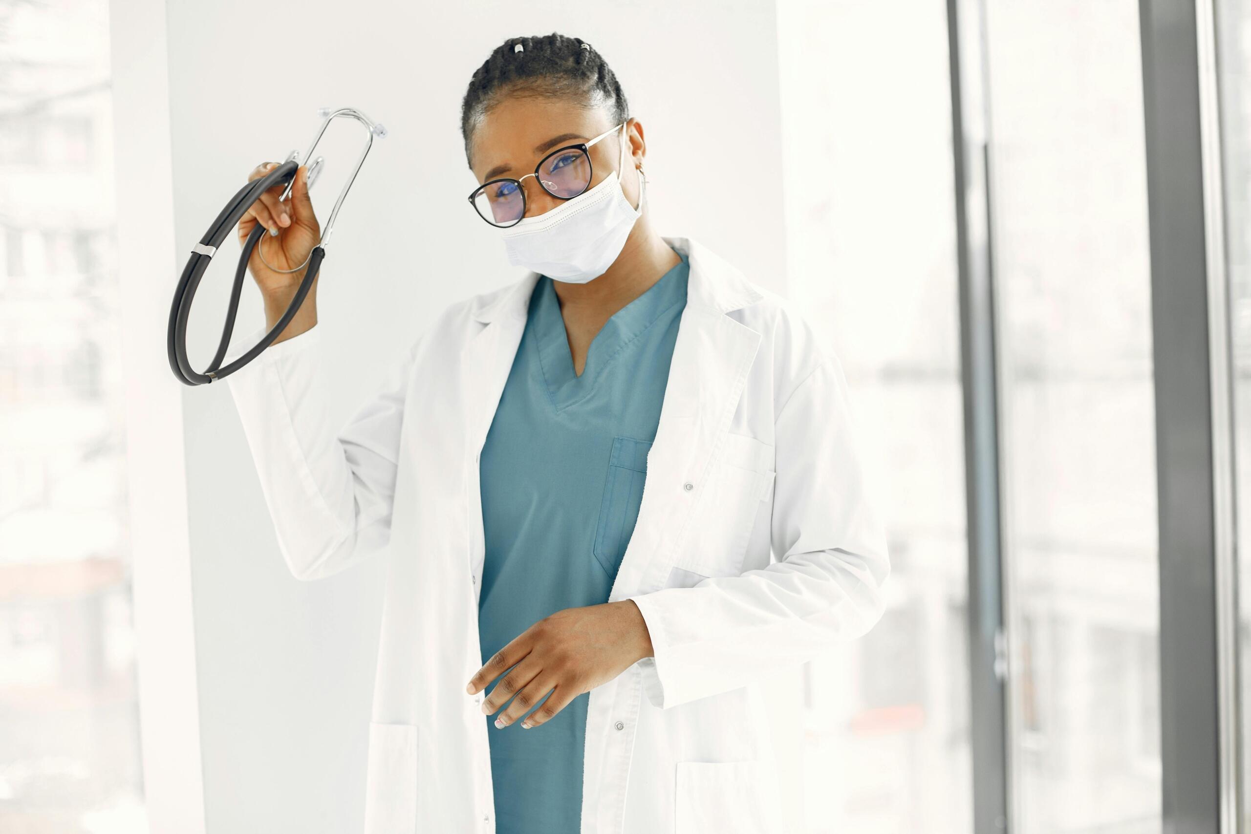 smiling doctor in lab coat, wearing a surgical mask with stethoscope in her hand