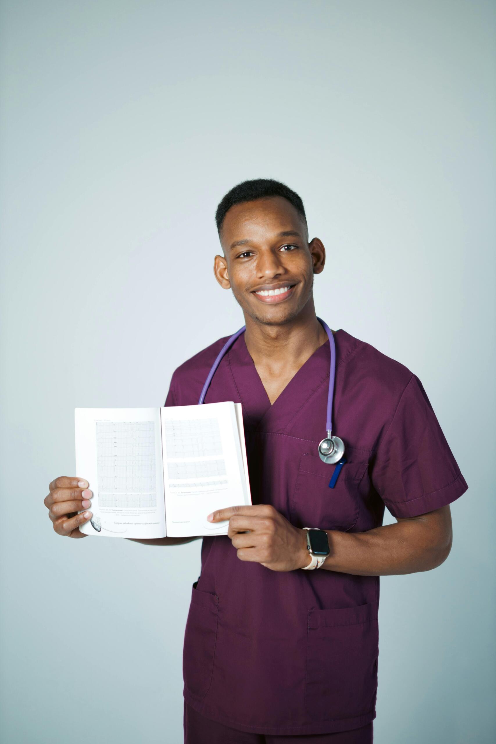 smiling doctor in burgundy scrubs with stethoscope around his neck and holding up an open book