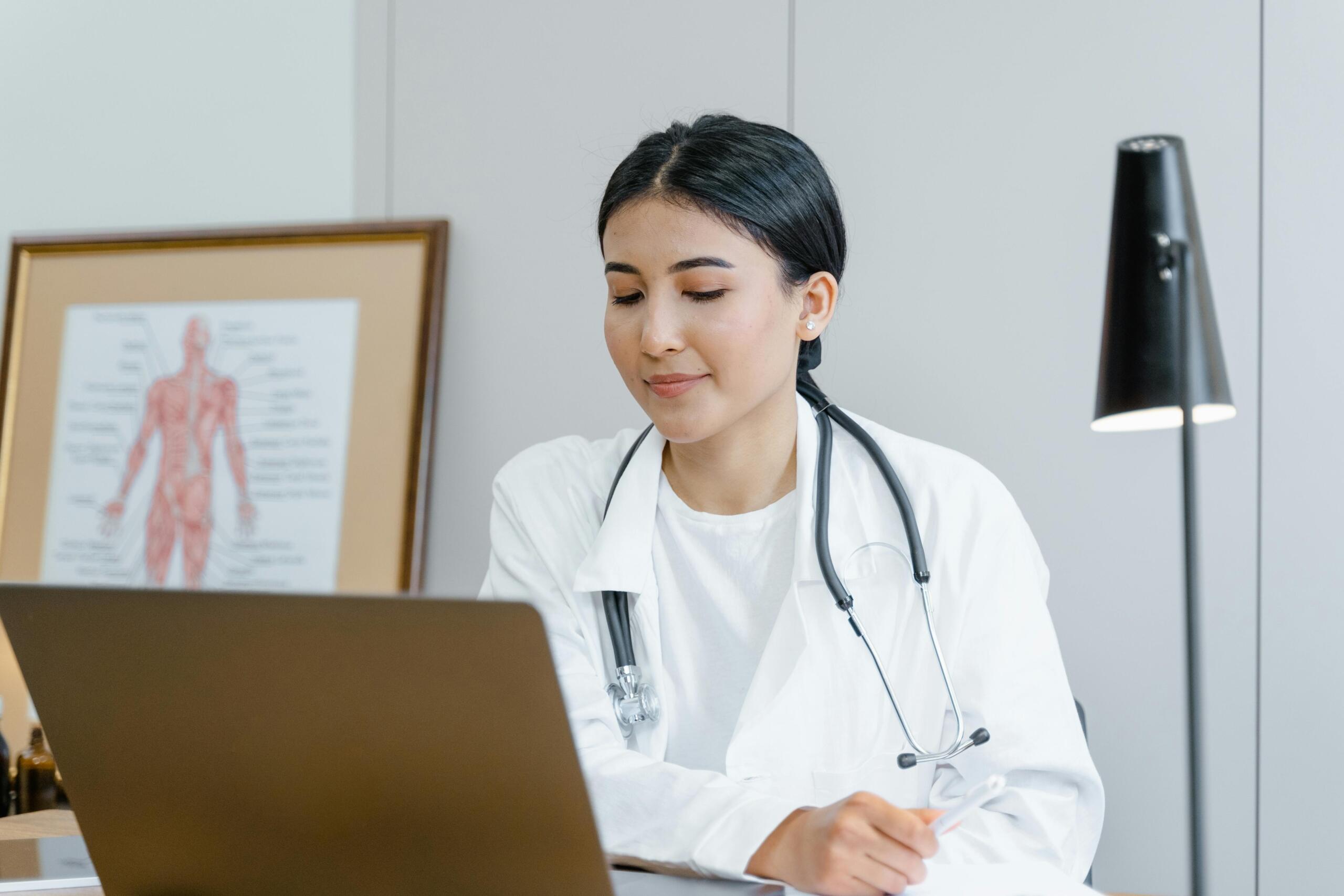 doctor in white lab coat with stethoscope around her neck sitting behind a desk using laptop