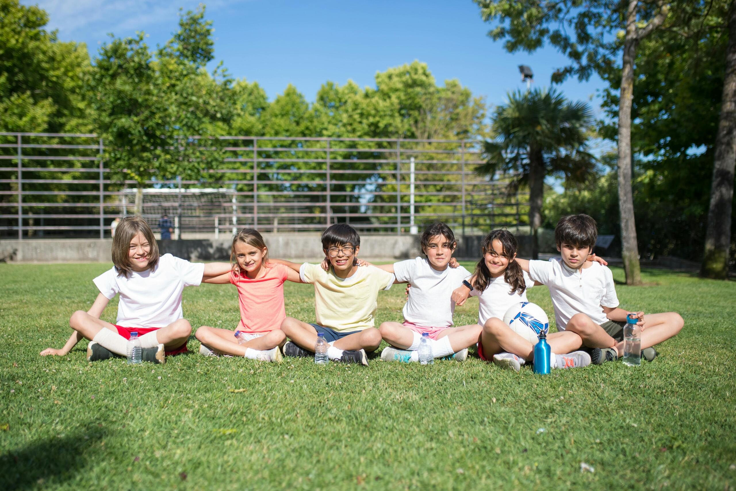 young children sitting side by side on grass with their arms around each others' shoulders