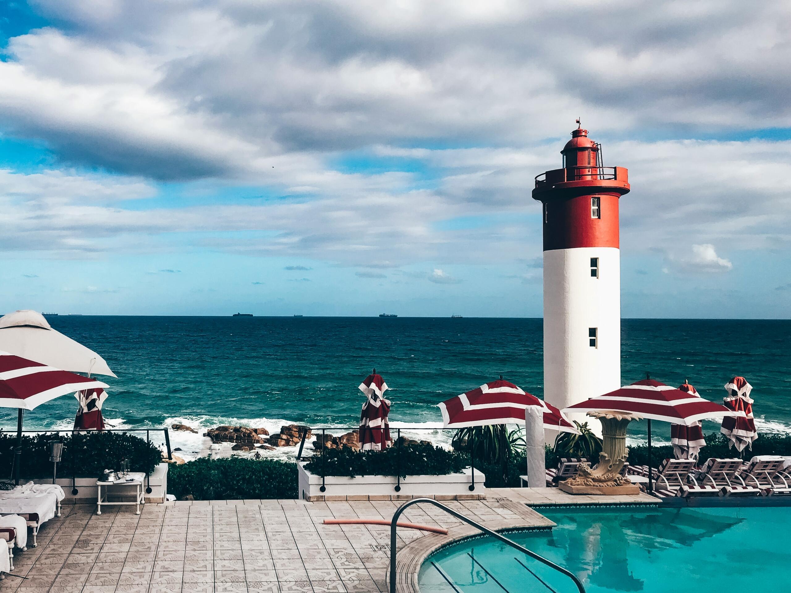 red and white lighthous on a rock shore in front of red and white umbrellas and a swimming pool