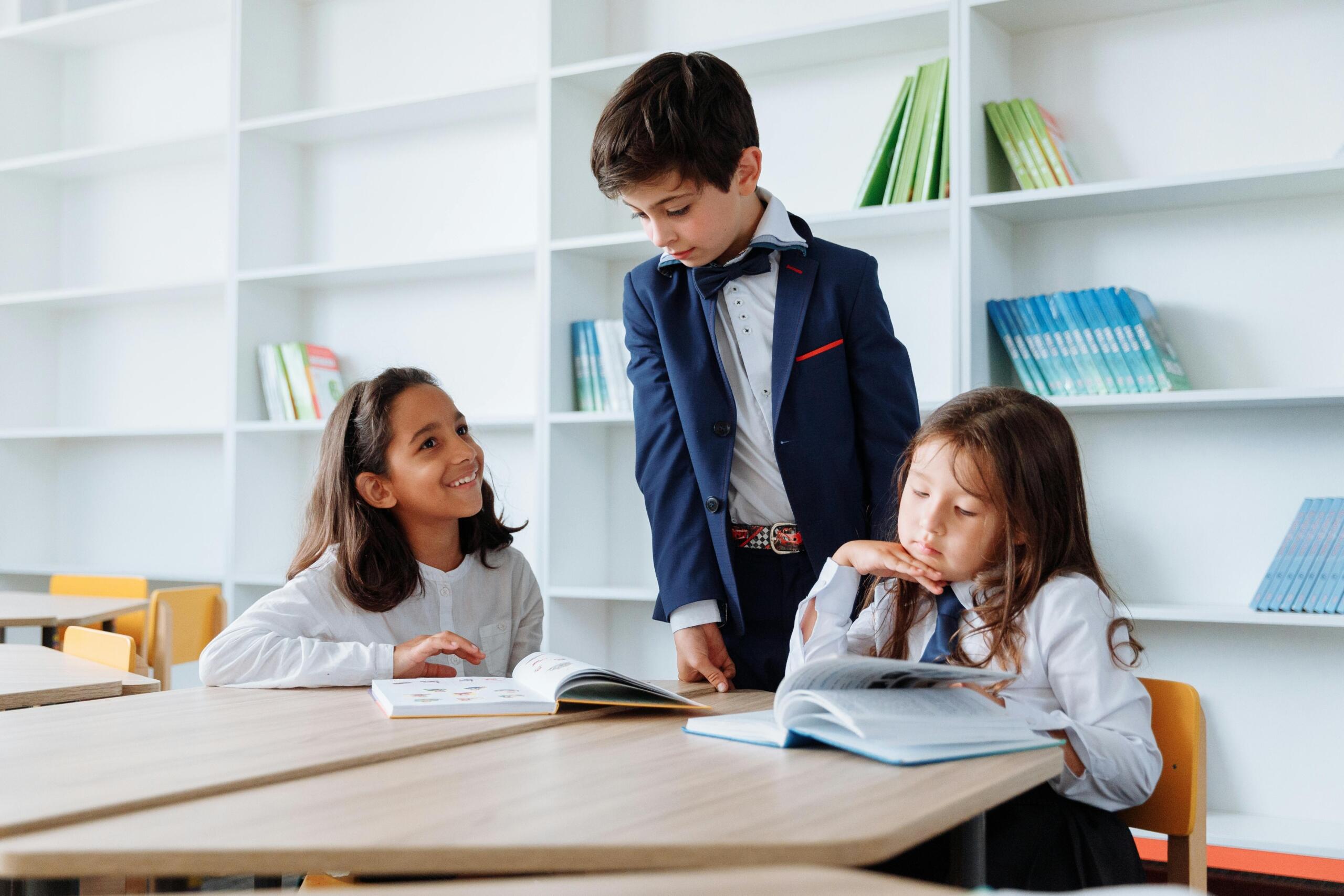 two school girls sitting at a table reading while a school boy looks on