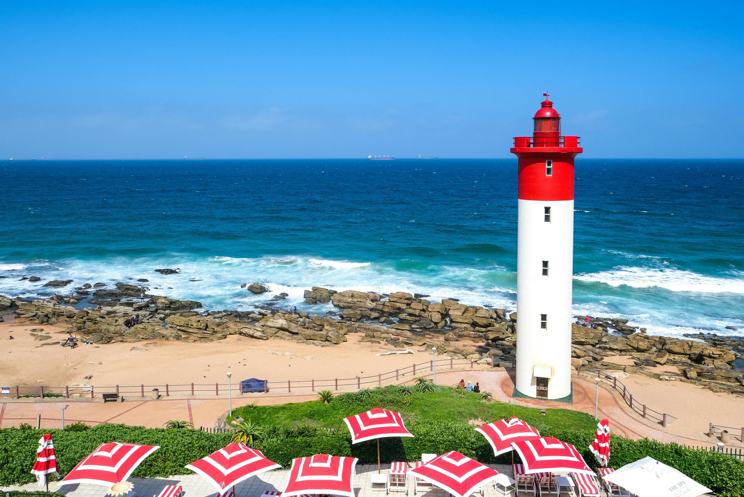 red and white lighthous on a rock shore in front of red and white umbrellas