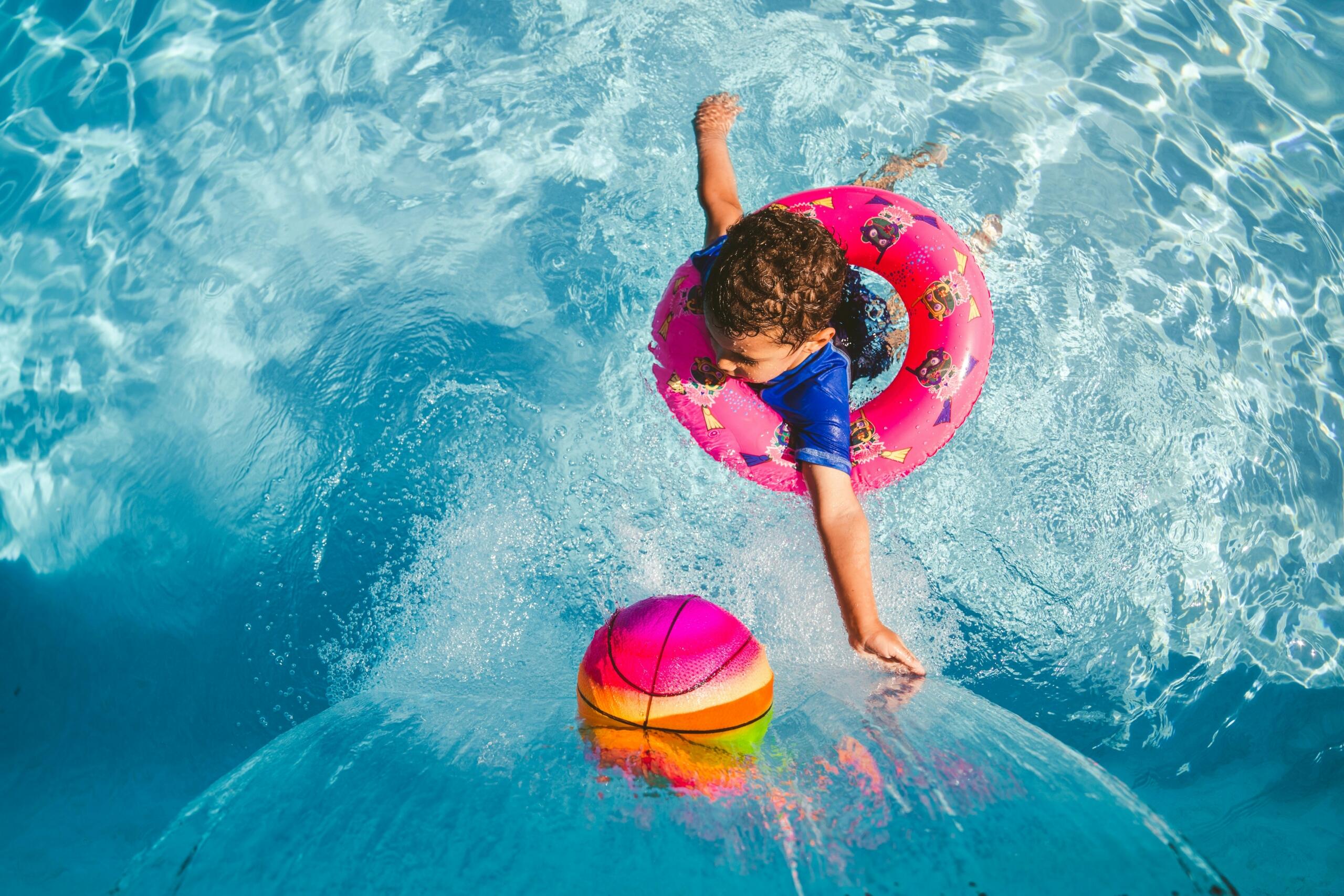 young child in a swimming pool with a water feature playing with an inflatable ball and using a swimming tube