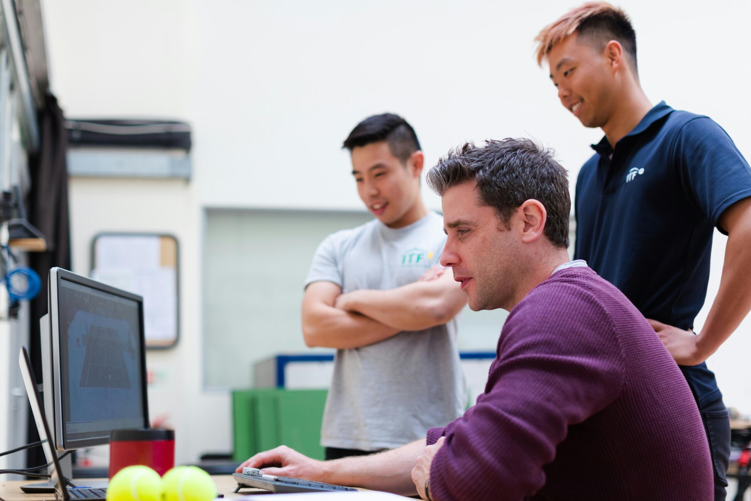 man-in-maroon-long-sleeve-shirt-working a on desktop computer while his colleagues look at his screen