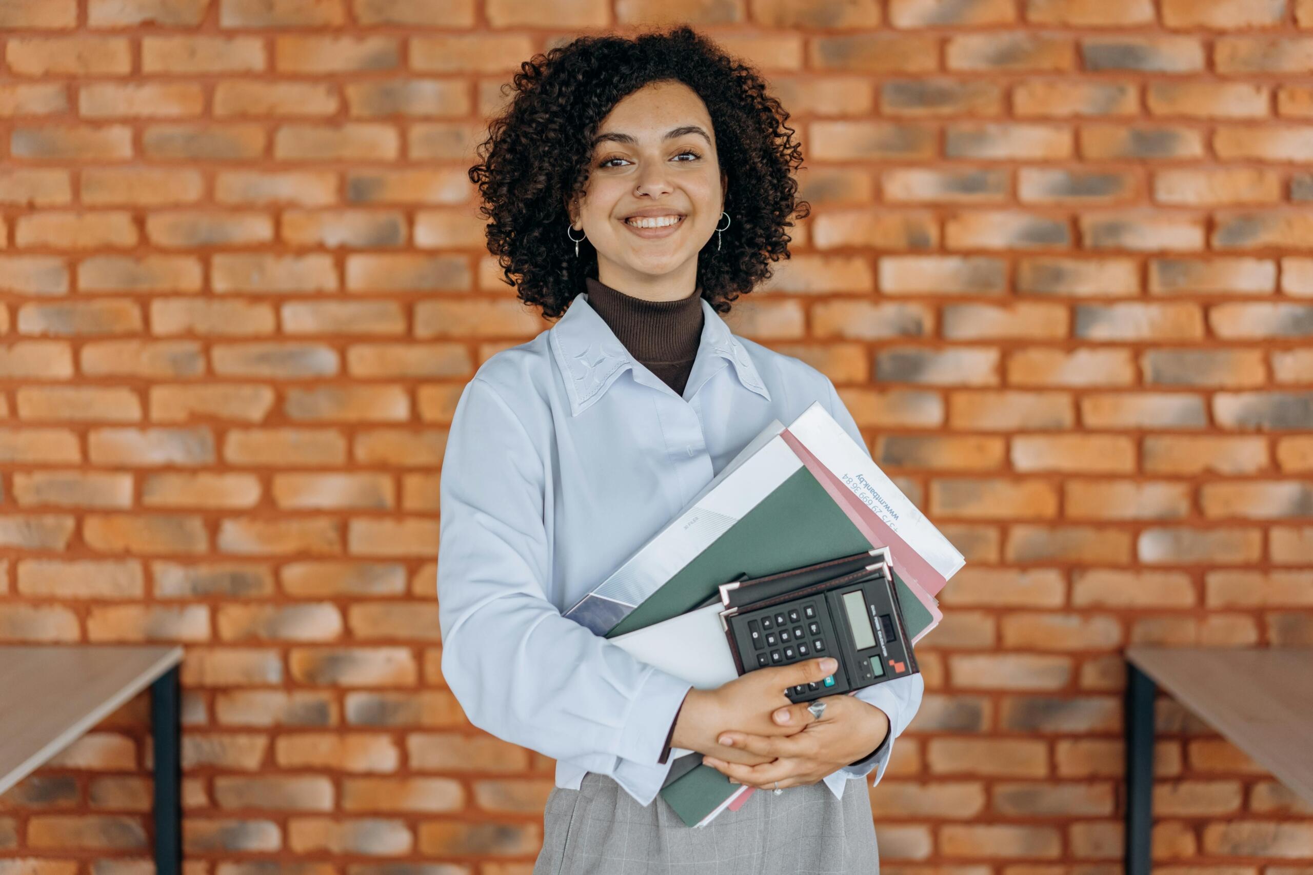 smiling woman in smart casual attire holding notebooks and a calculator