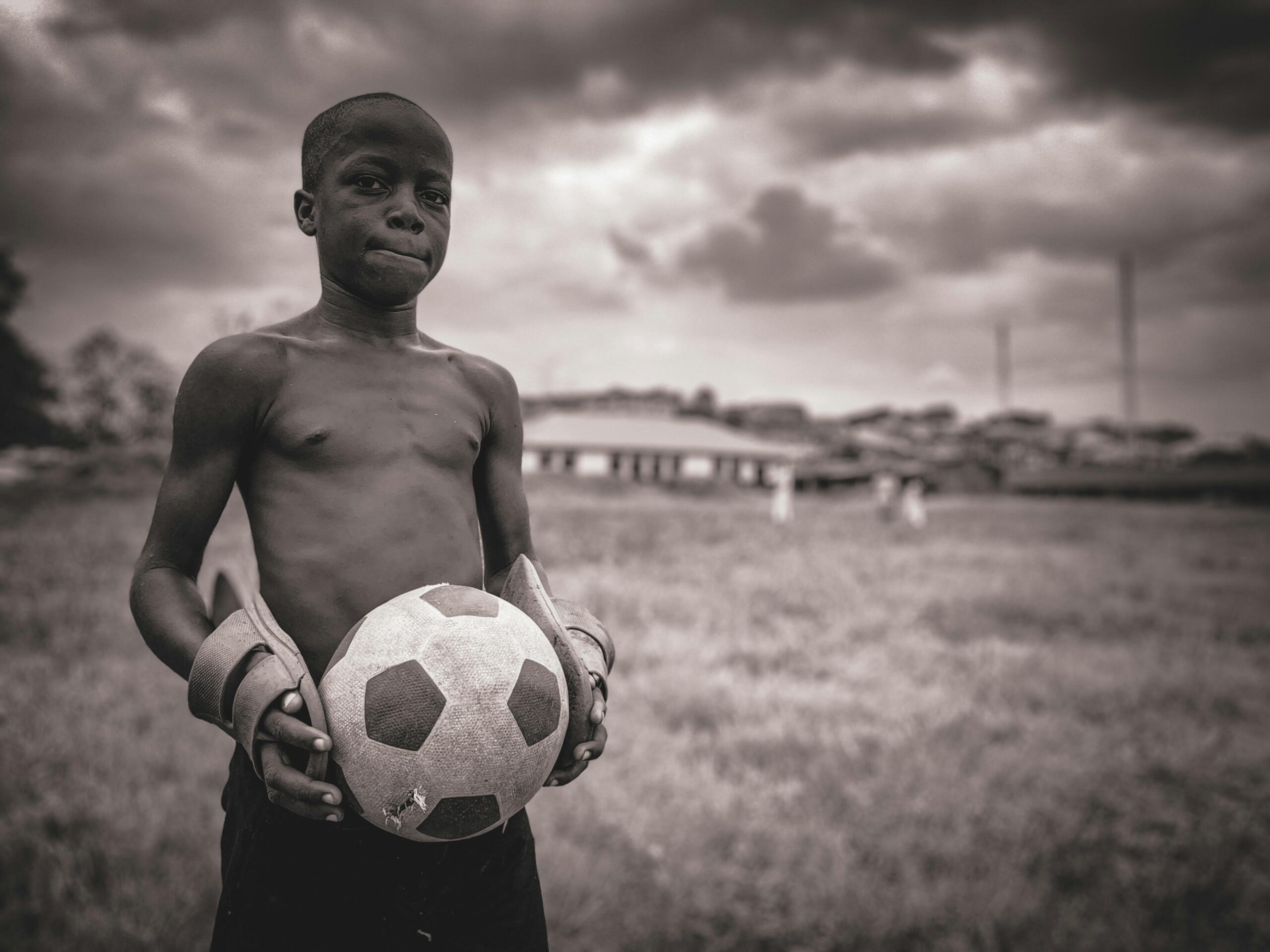 A black and white image of a young boy holding a ball on a soccer pitch.