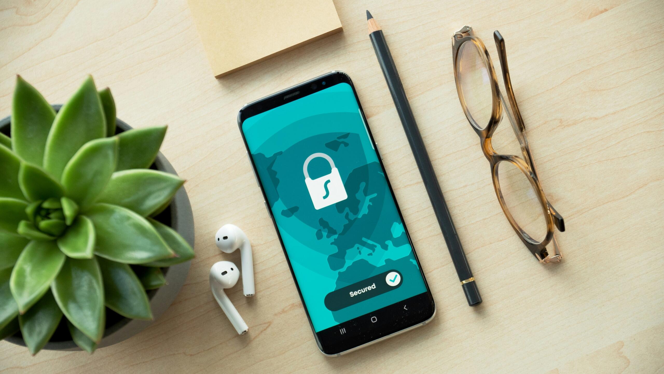 Phone with a lock on it on a desk with earphones, a pencil, glasses, a notepad, and a plant.