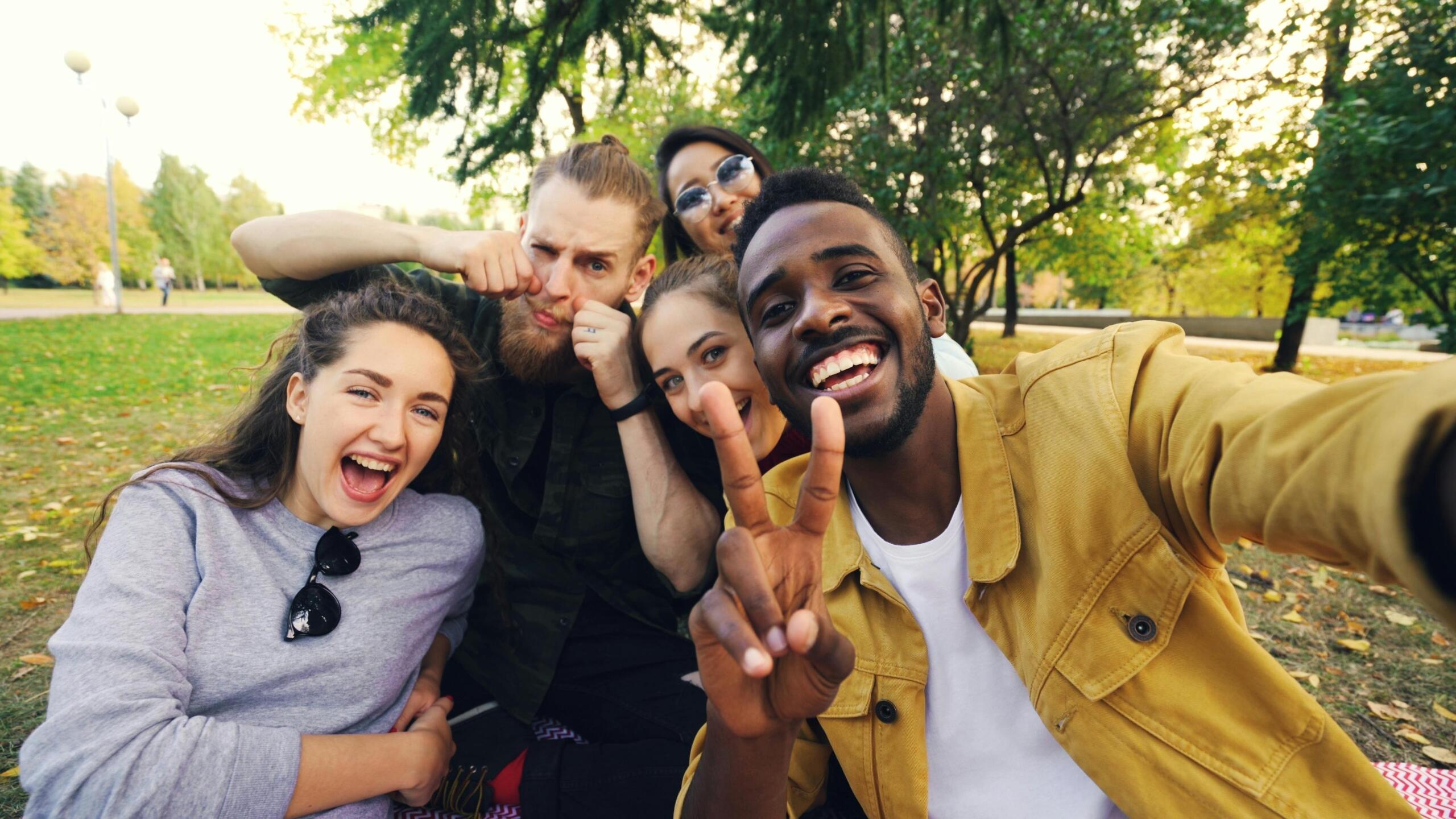 A diverse group of smiling friends taking a selfie under trees during the day.