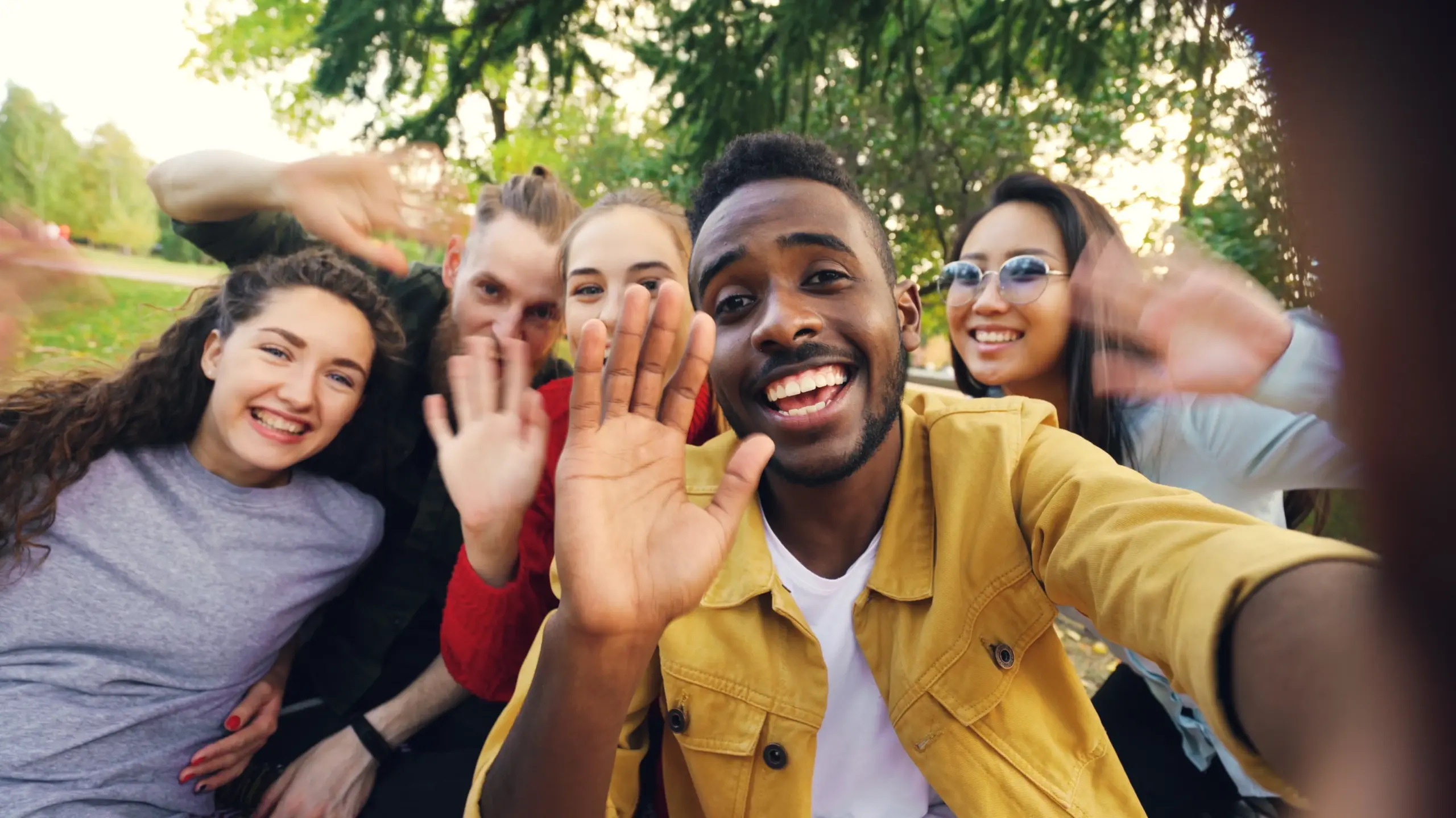 A diverse group of smiling friends taking a selfie under trees during the day.