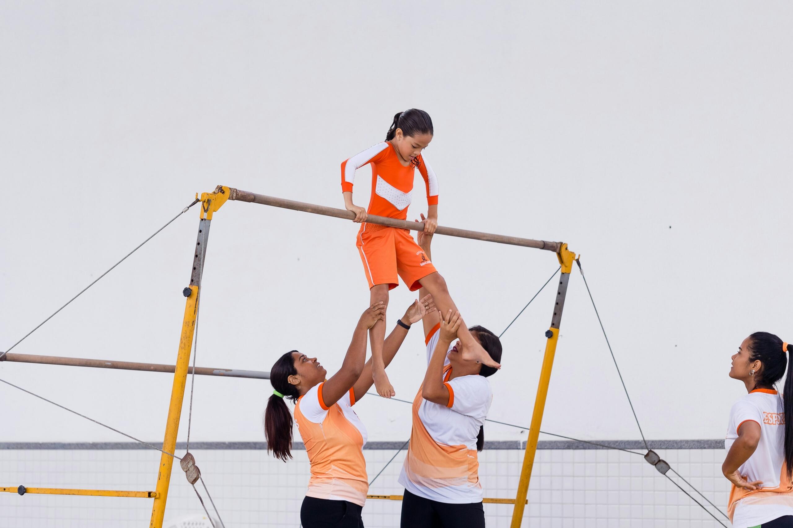 Two coaches assist a young gymnast to hold onto bars.