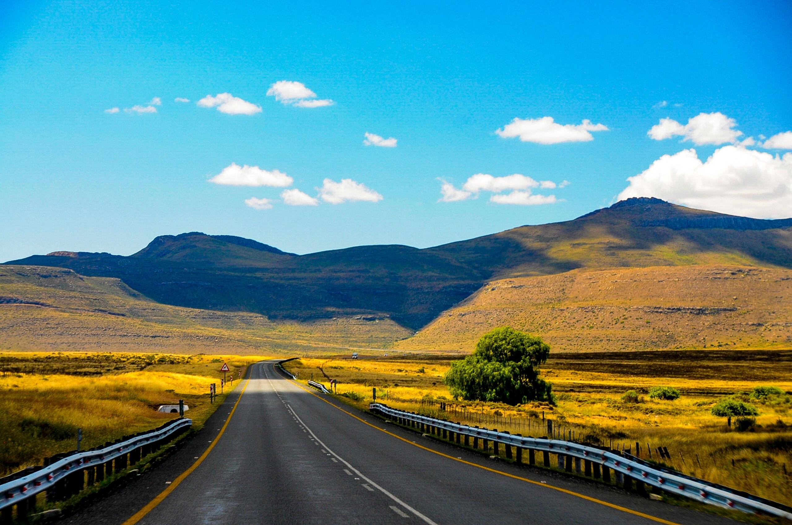 A scenic open road stretches through mountains and open fields under a bright blue sky.