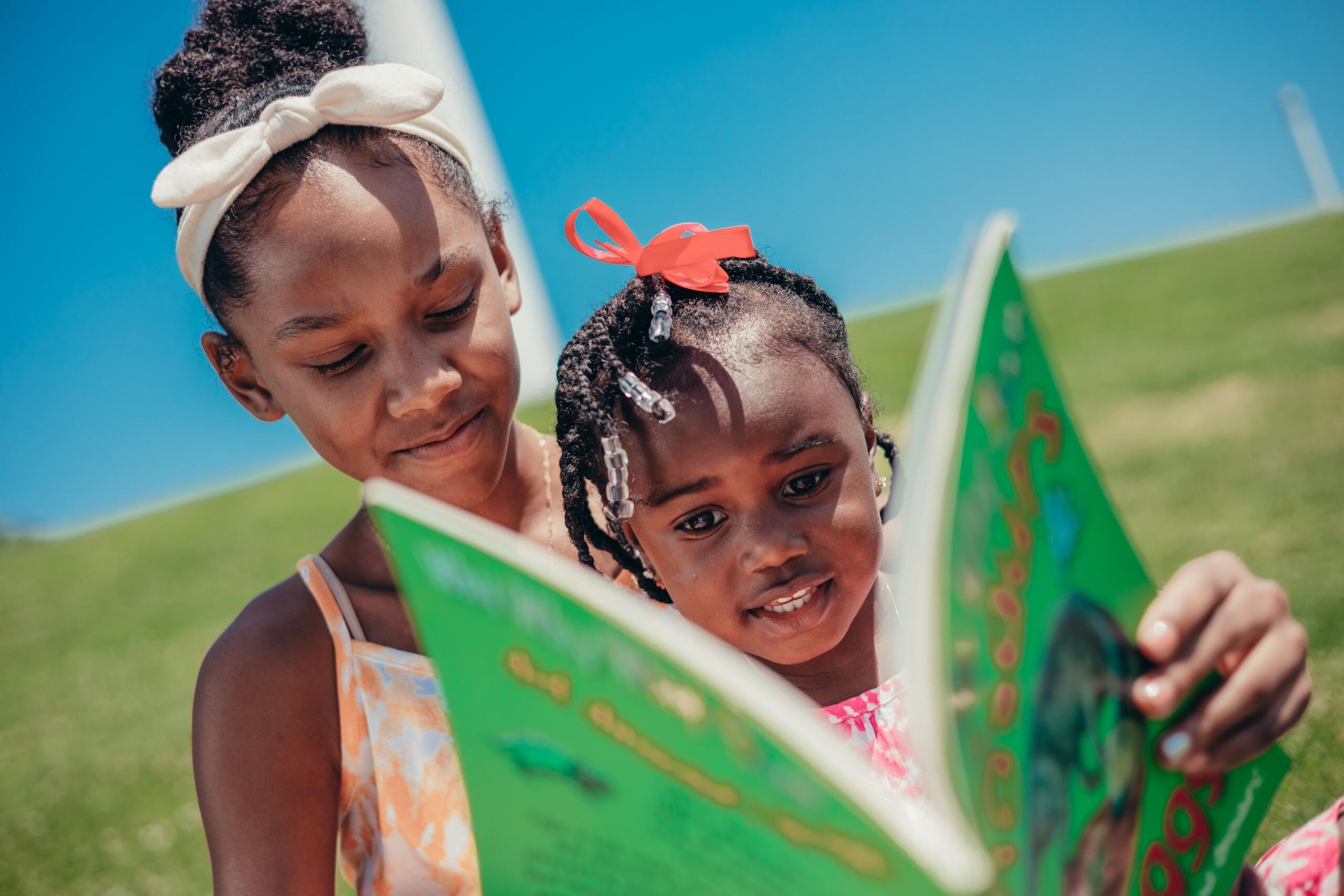 two young girls sitting outside on grass reading a book