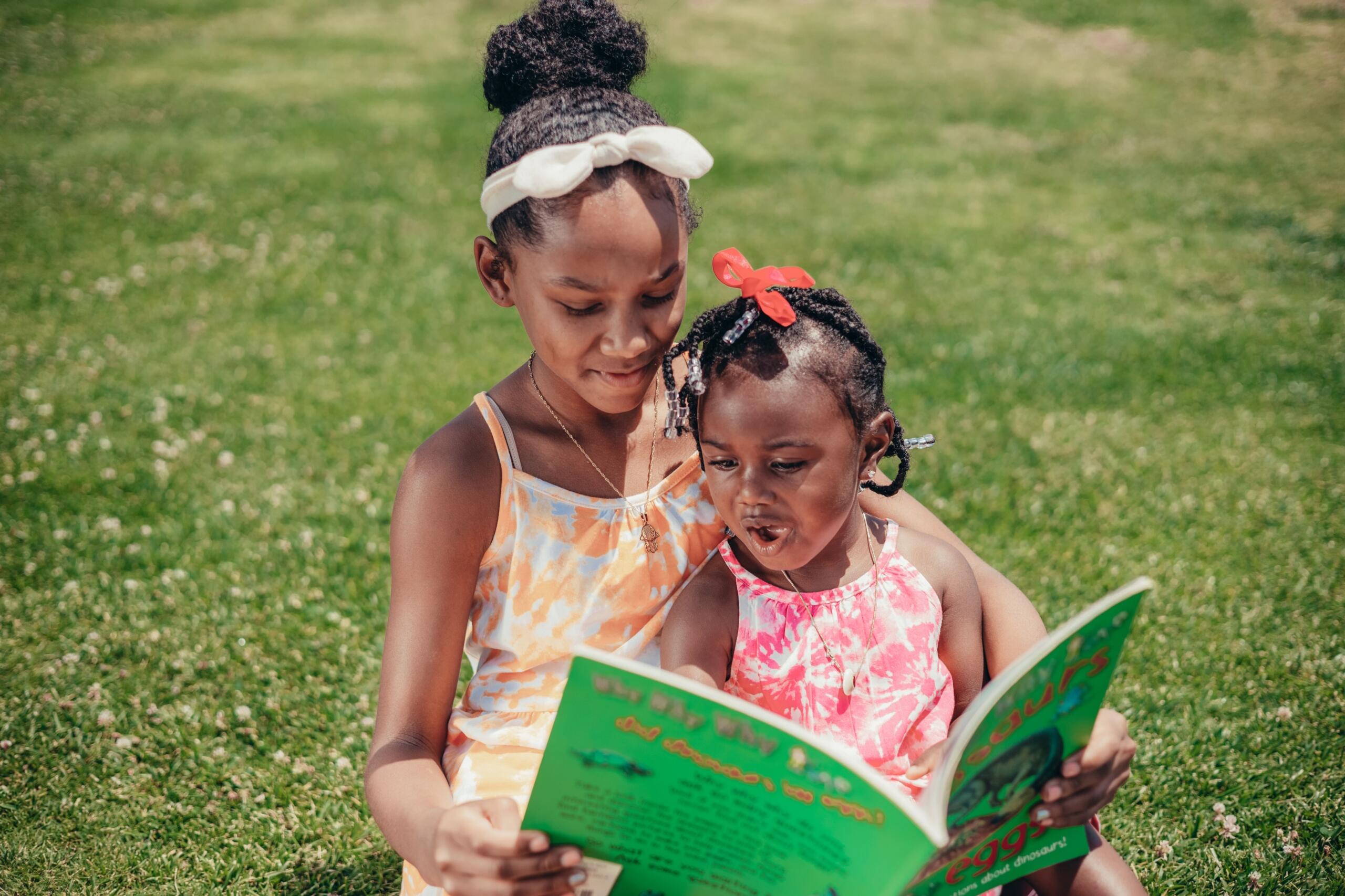 two young girls sitting outside on grass reading a book