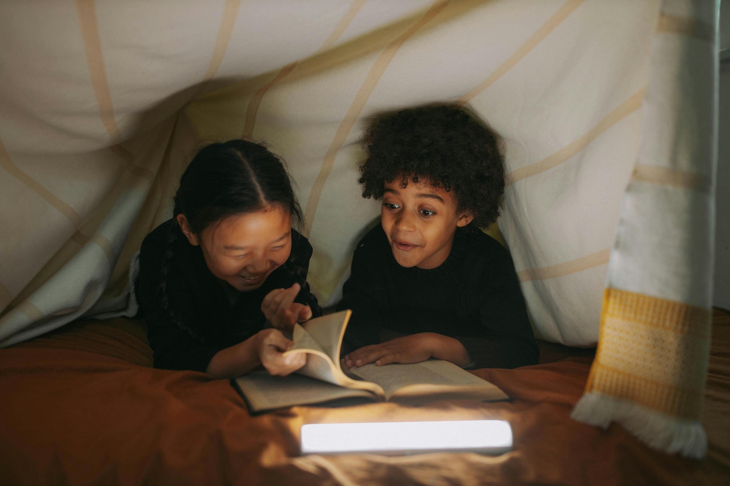 two laughing children in an indoor tent reading a book by torchlight