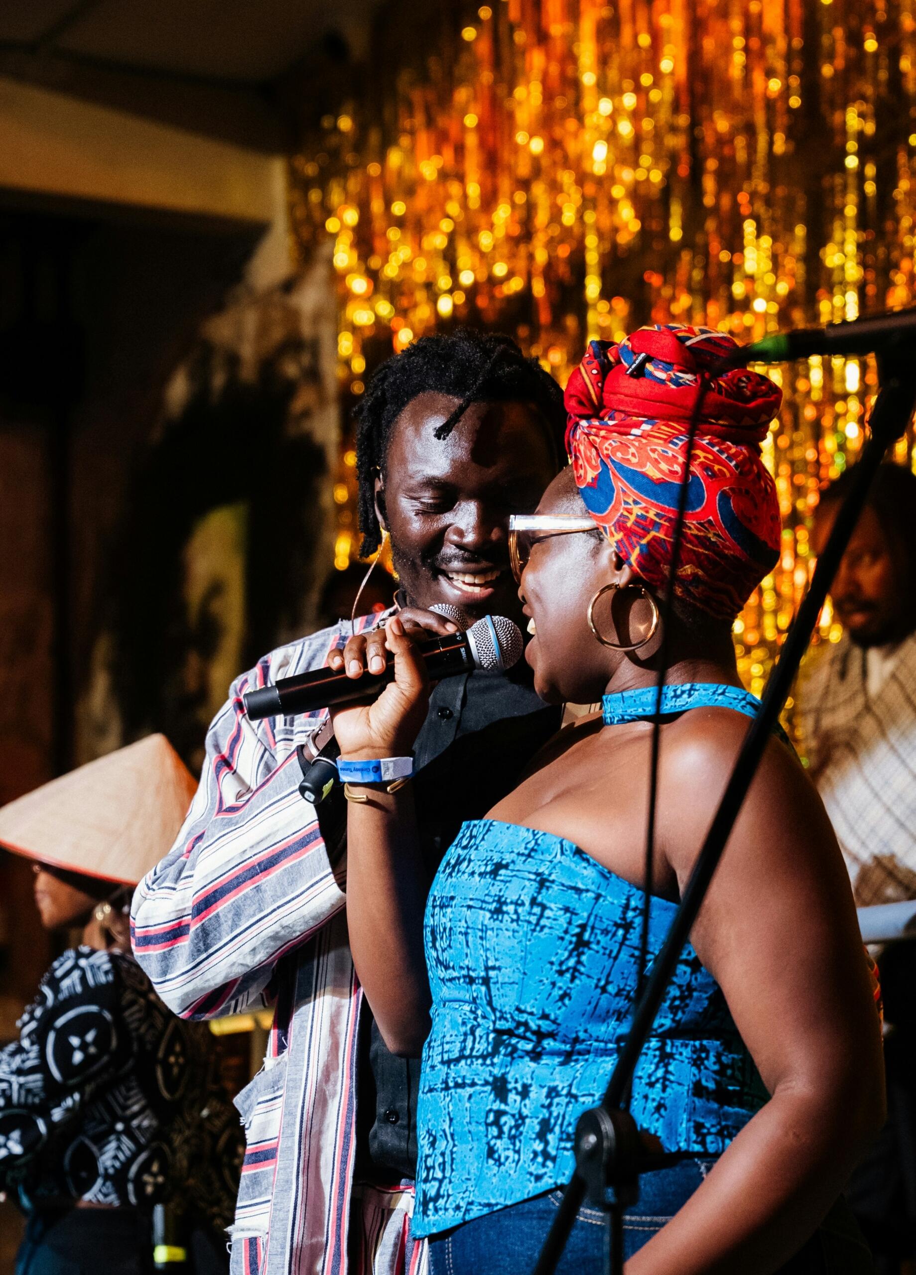 African singers performing together on stage, with a female singer in colourful traditional clothing singing into a microphone beside a male singer.