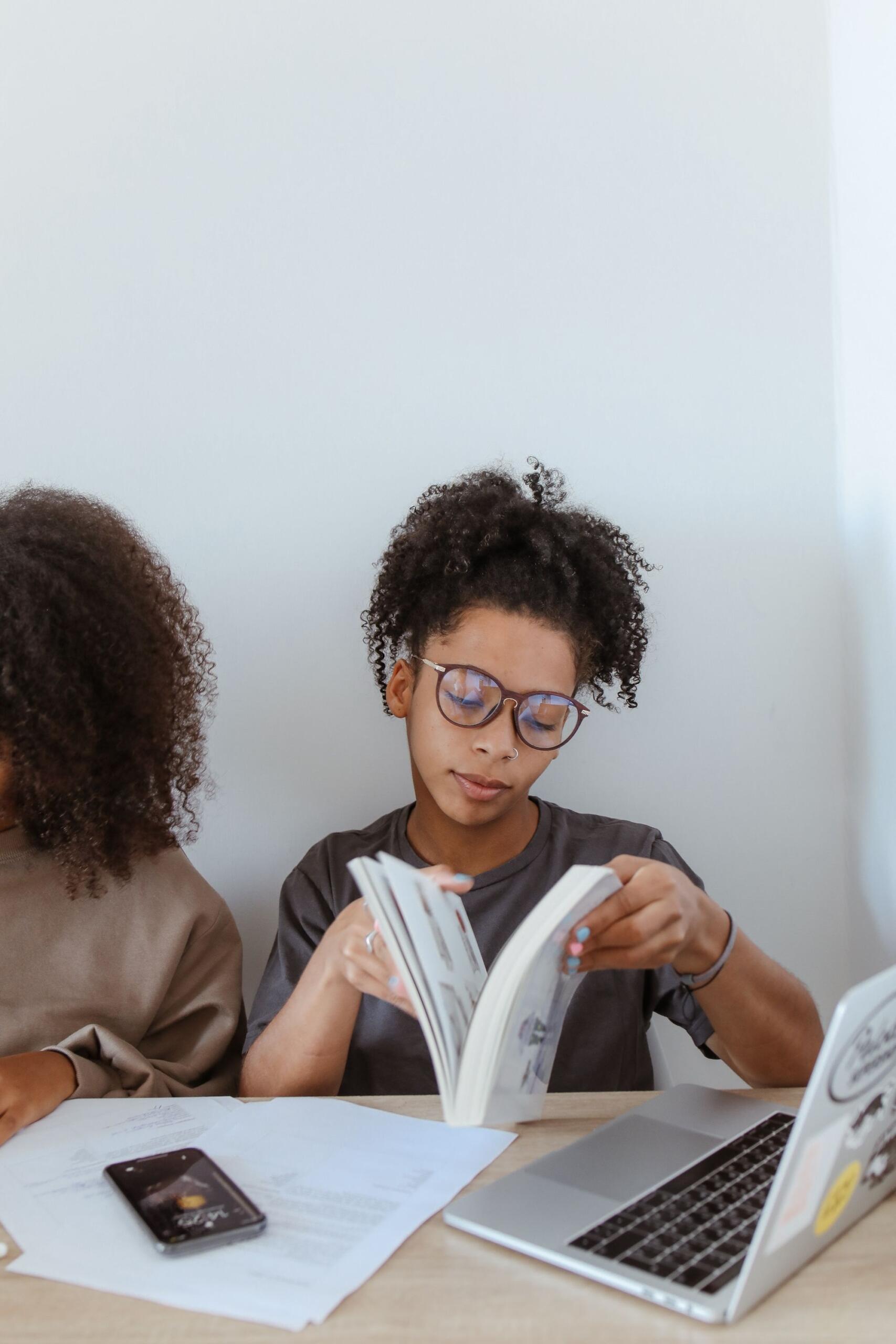 bespectacled teen opening a book beside a laptop
