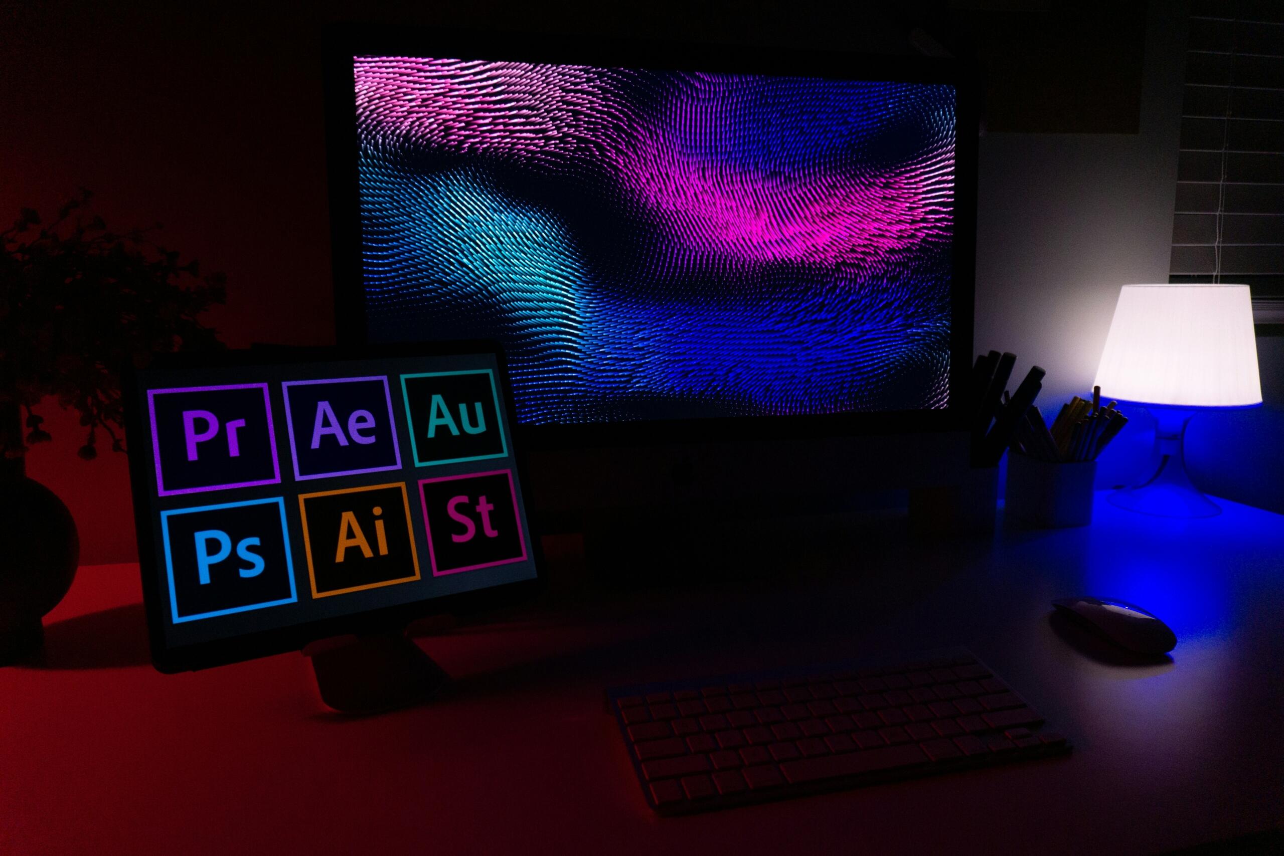 Dual monitor desk setup at night showing creative software app icons and a colourful abstract wave pattern
