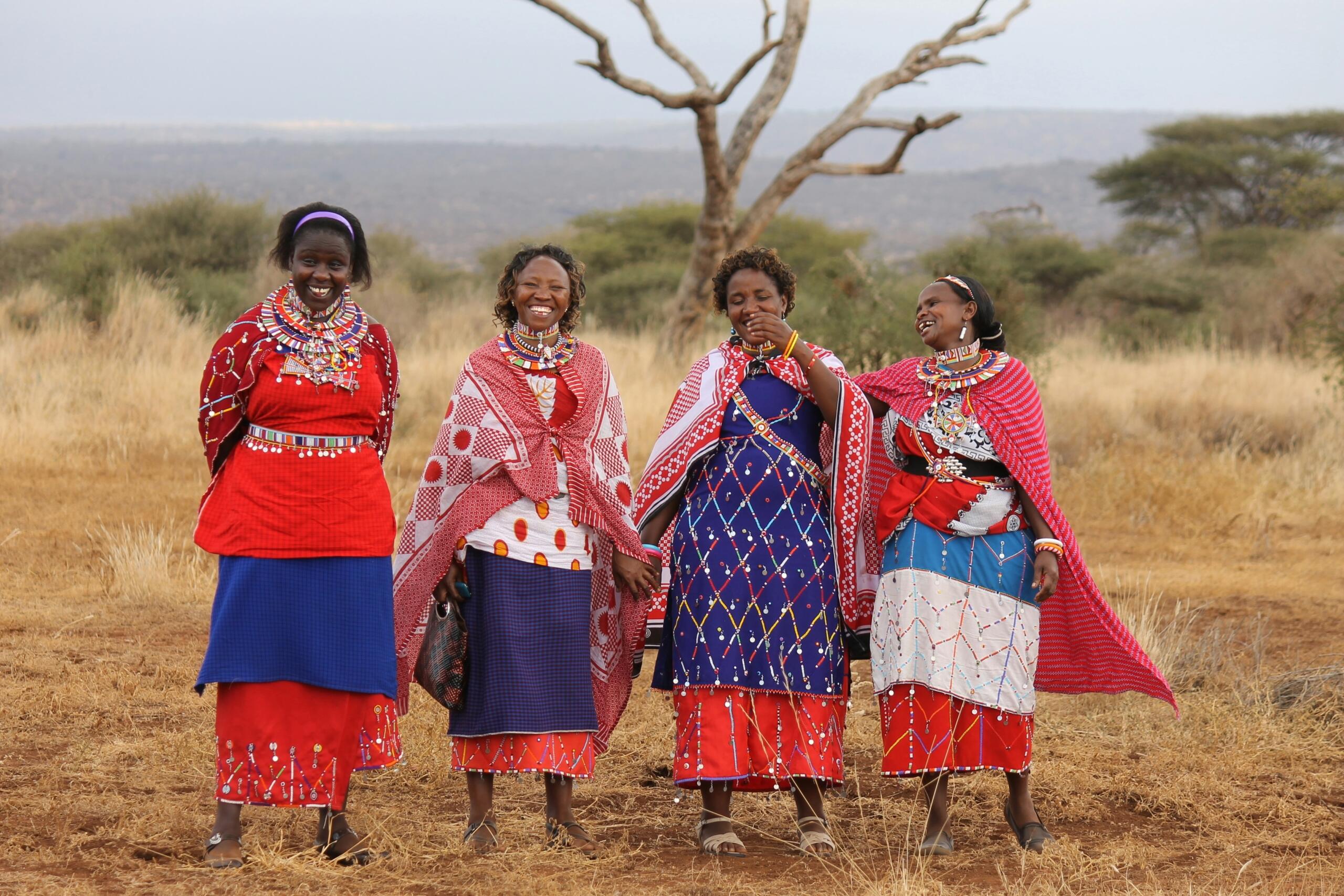 Four Maasai women standing together in colourful traditional clothing in a grassland landscape.