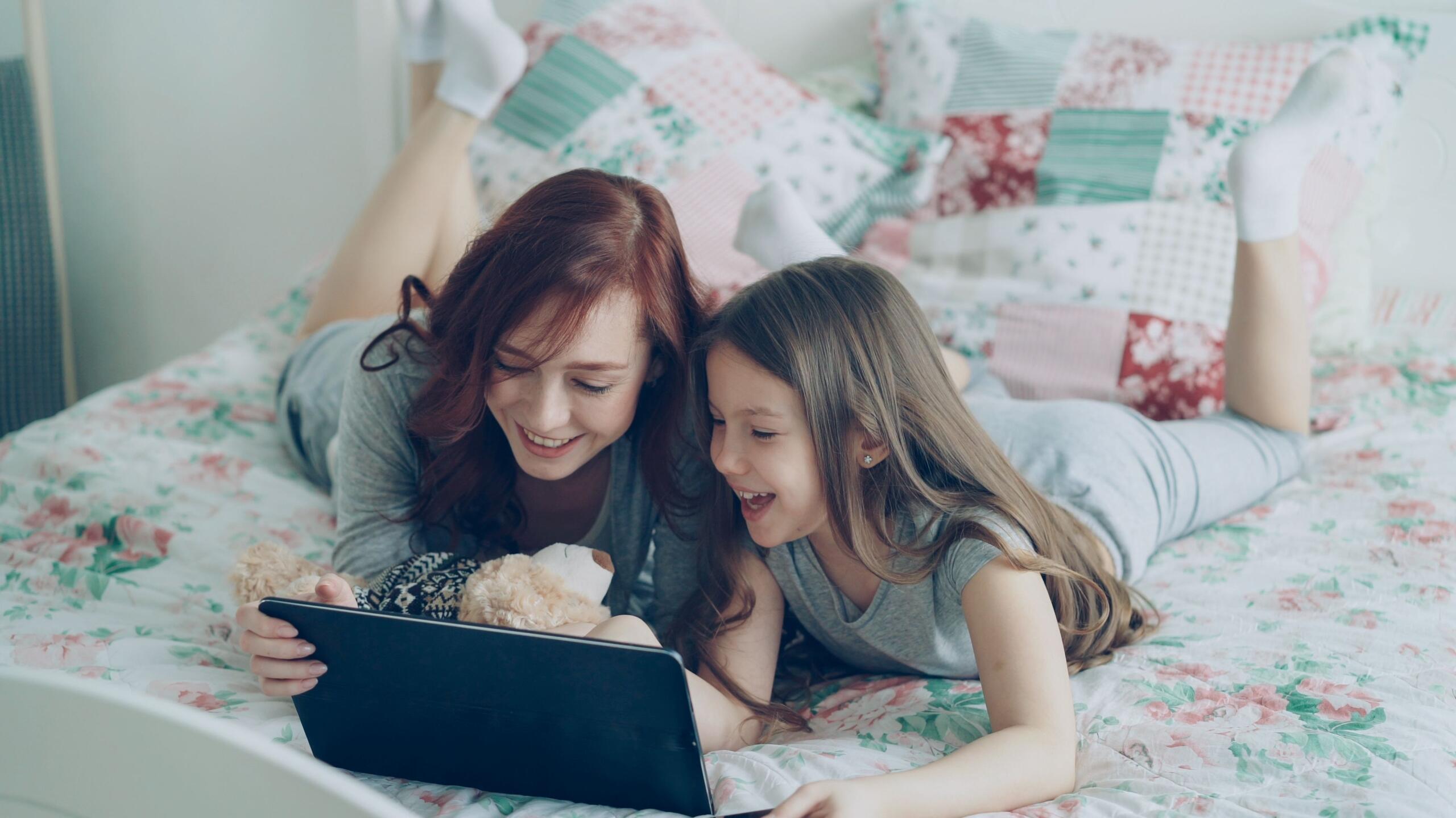 mother and child lying on a bed reading an e-reader together