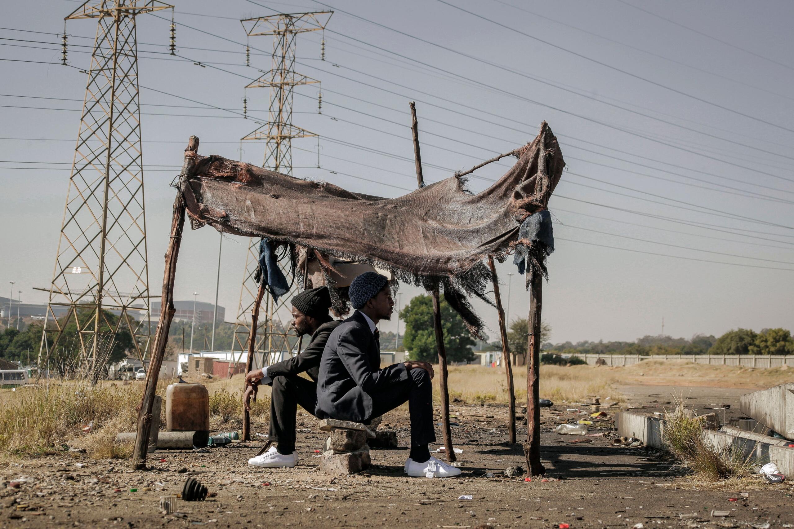 People sitting beside a makeshift shelter in Soweto, Johannesburg, with the urban landscape visible behind them.