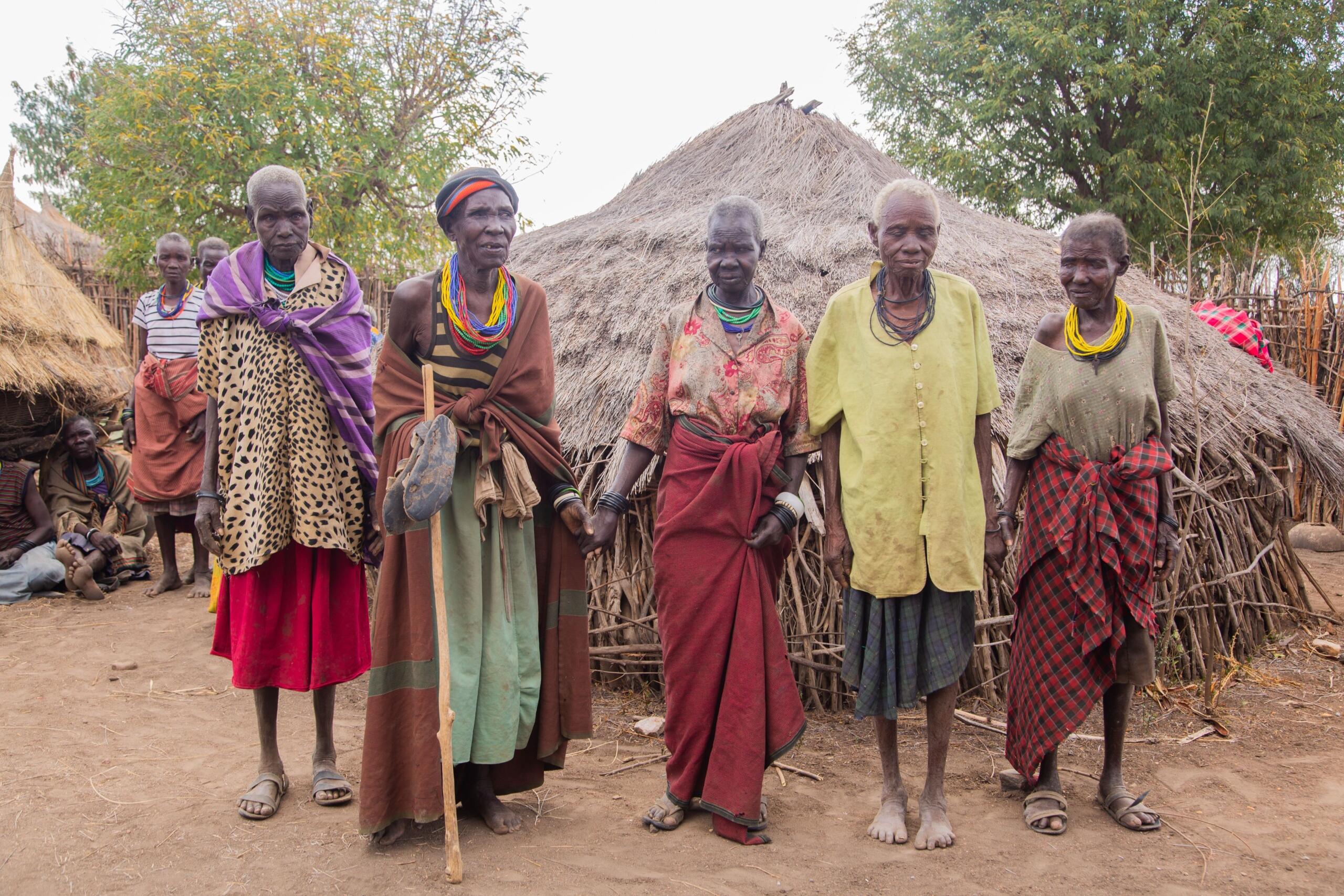 Group of African elders standing outside traditional thatched homes, wearing colourful clothing and beadwork.