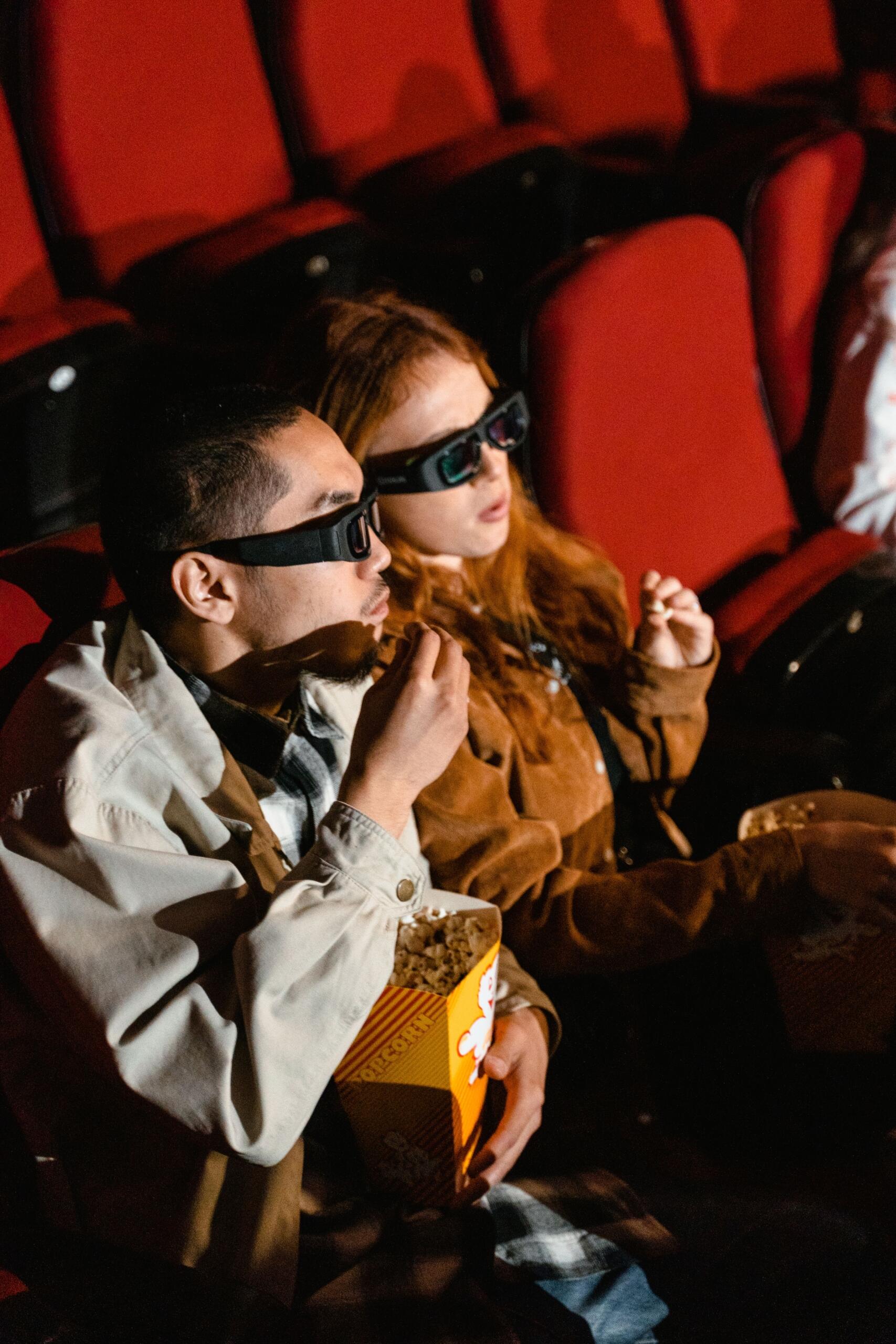 Two teens are engrossed in a film and eating popcorn at the cinema.