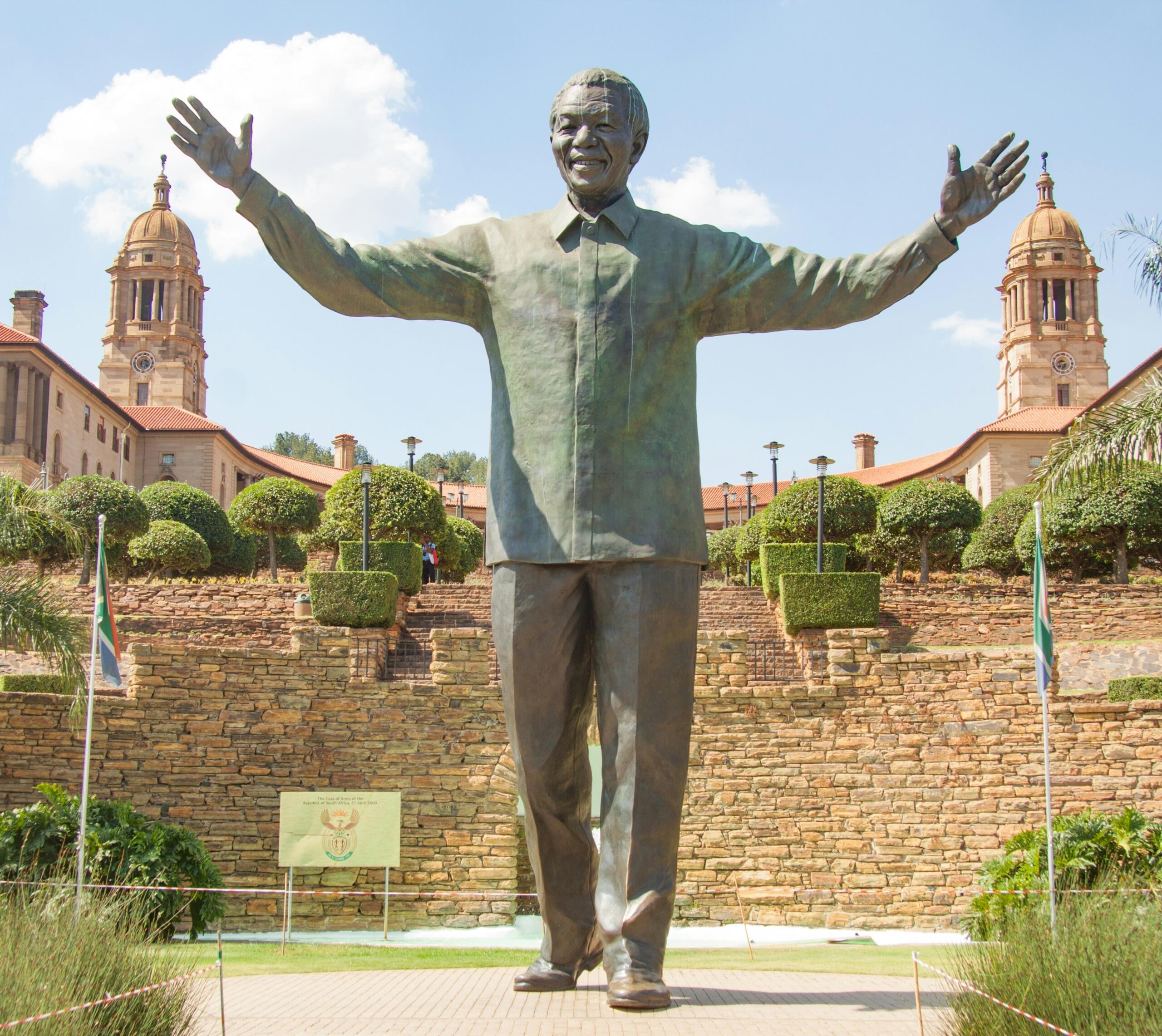 Nelson Mandela statue at the Union Buildings in Pretoria, with the South African flag and historic government buildings behind it.
