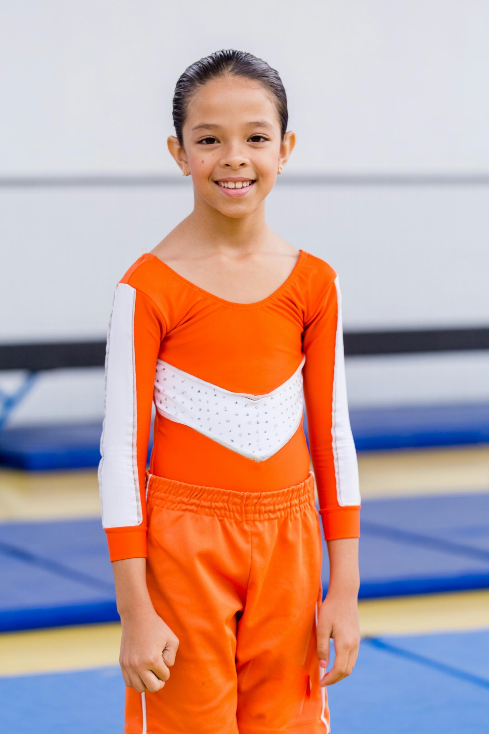 smiling girl wearing long-sleeved orange and white gymnastics leotard and tracksuit bottoms